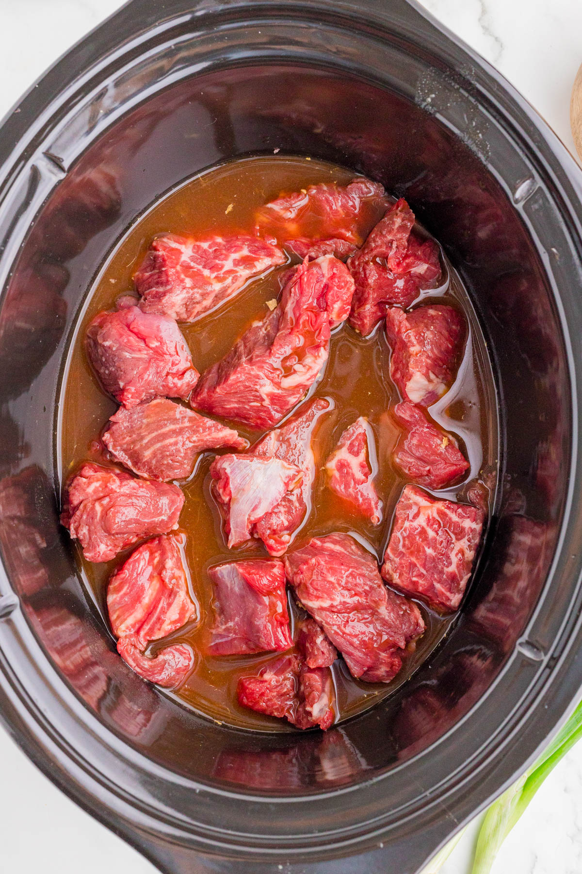 Chunks of raw beef sitting in brown liquid inside a black slow cooker, ready to be cooked.