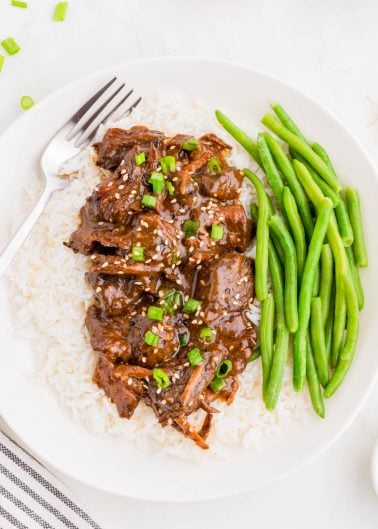 A plate of white rice topped with shredded beef in a dark sauce, garnished with green onions and sesame seeds, served with green beans and a fork on the side.