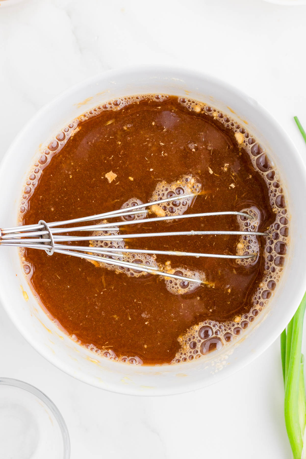 A metal whisk rests in a white bowl filled with a brown liquid mixture.