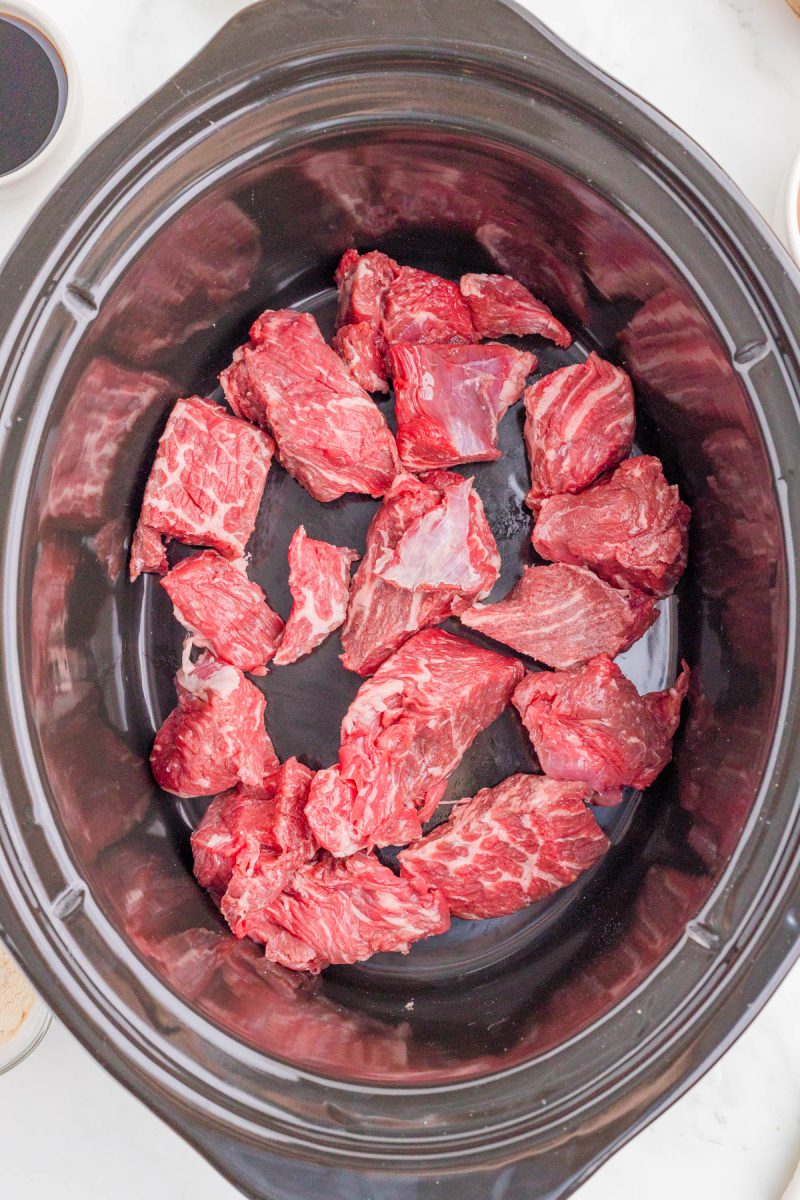 Chunks of raw beef placed in the bottom of a black slow cooker.