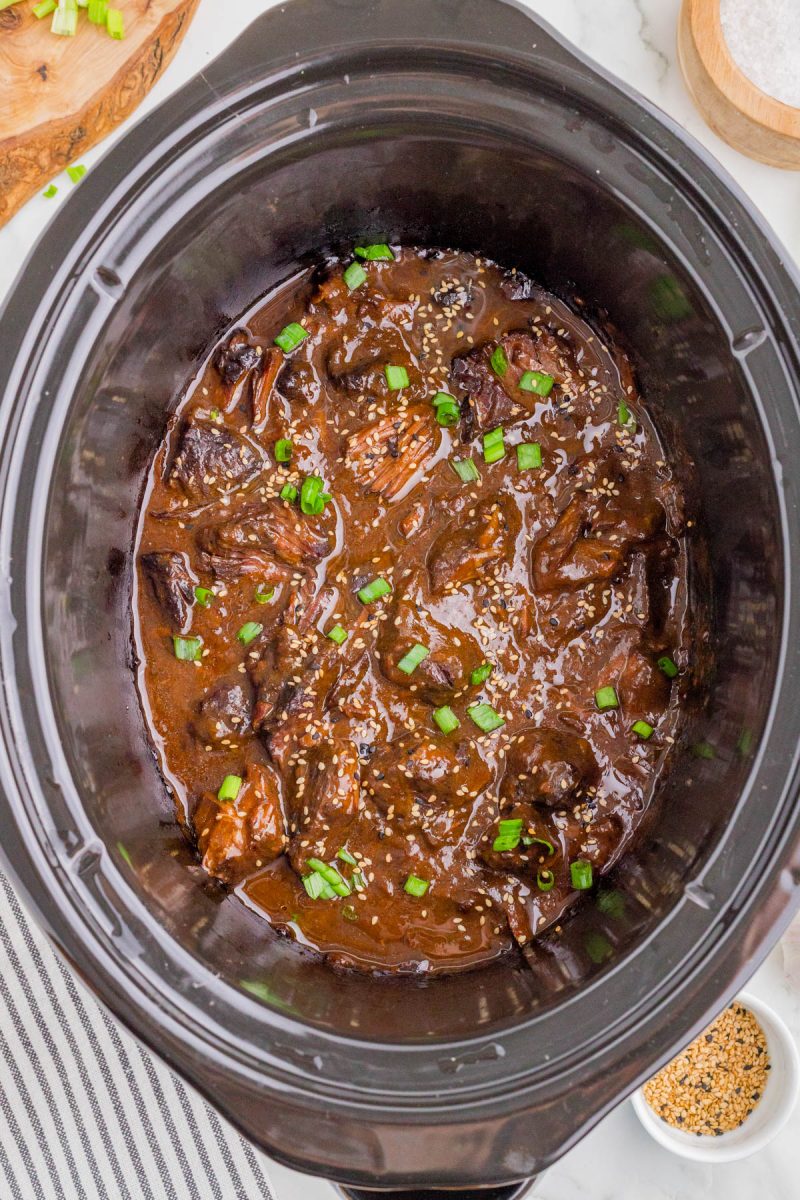 Shredded beef in a dark sauce garnished with chopped green onions and sesame seeds inside a black slow cooker.