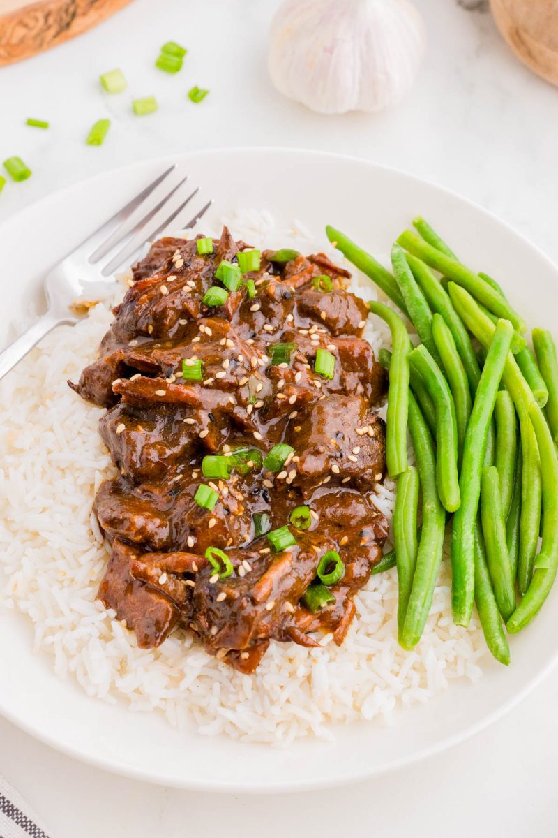A plate of white rice topped with shredded beef in brown sauce, garnished with green onions and sesame seeds, served with cooked green beans. A fork is placed on the plate.