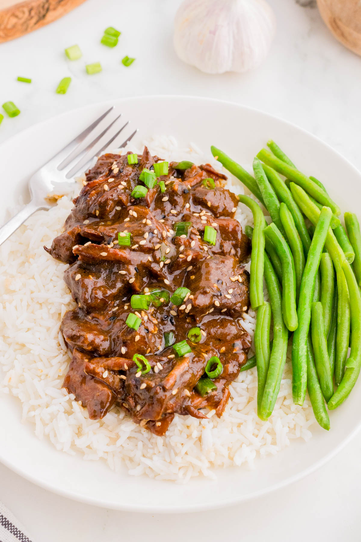 A plate of white rice topped with shredded beef in brown sauce, garnished with green onions and sesame seeds, served with cooked green beans. A fork is placed on the plate.