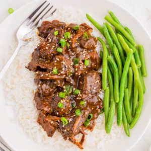 A plate of white rice topped with saucy beef, garnished with green onions and sesame seeds, served with steamed green beans and a fork.