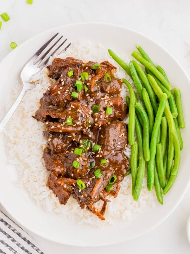 A plate of white rice topped with saucy beef, garnished with green onions and sesame seeds, served with steamed green beans and a fork.