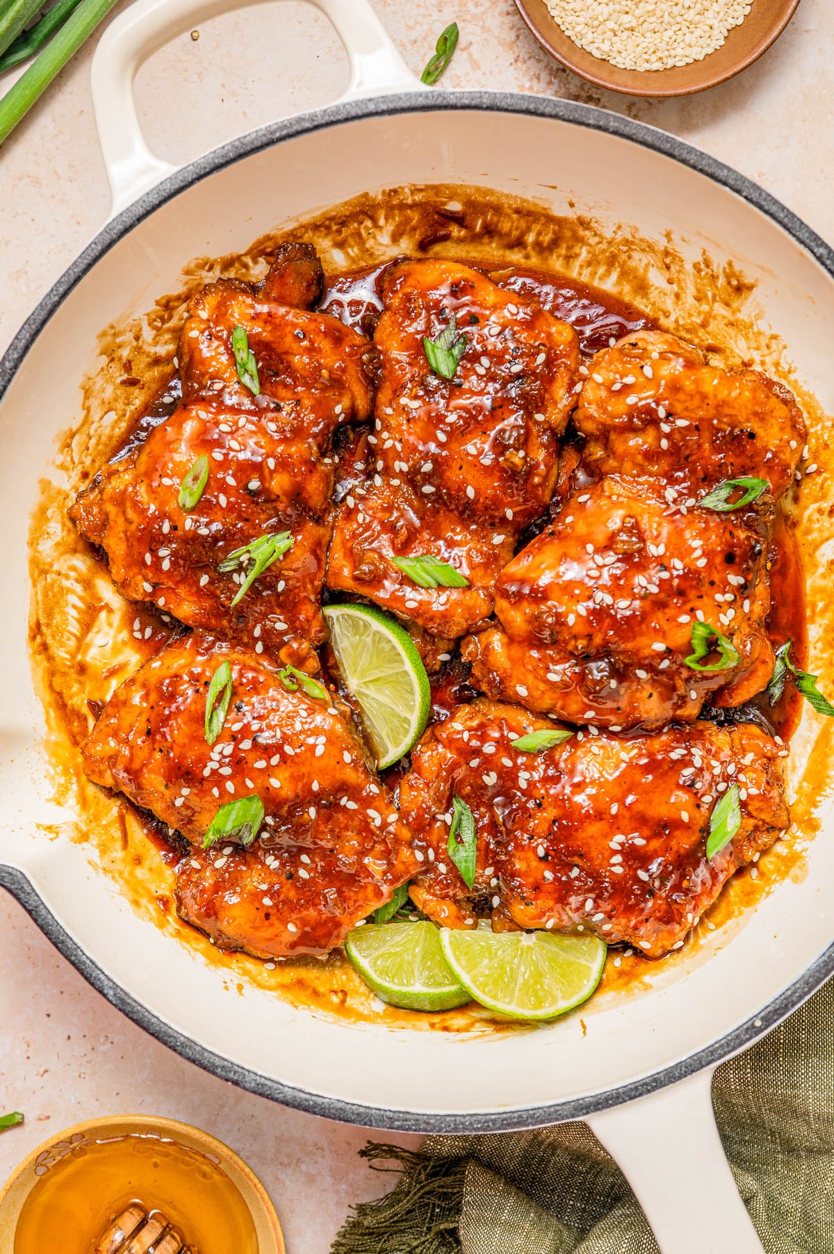 A skillet filled with glazed chicken thighs topped with sesame seeds and sliced green onions, garnished with lime wedges.
