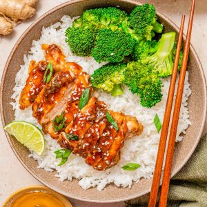 A bowl of white rice topped with glazed chicken, broccoli florets, sliced green onions, sesame seeds, and a lime wedge, with chopsticks resting on the side.