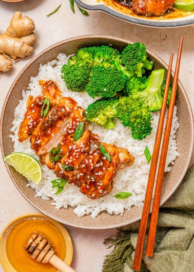 A bowl of white rice topped with glazed chicken, broccoli florets, sliced green onions, sesame seeds, and a lime wedge, with chopsticks resting on the side.