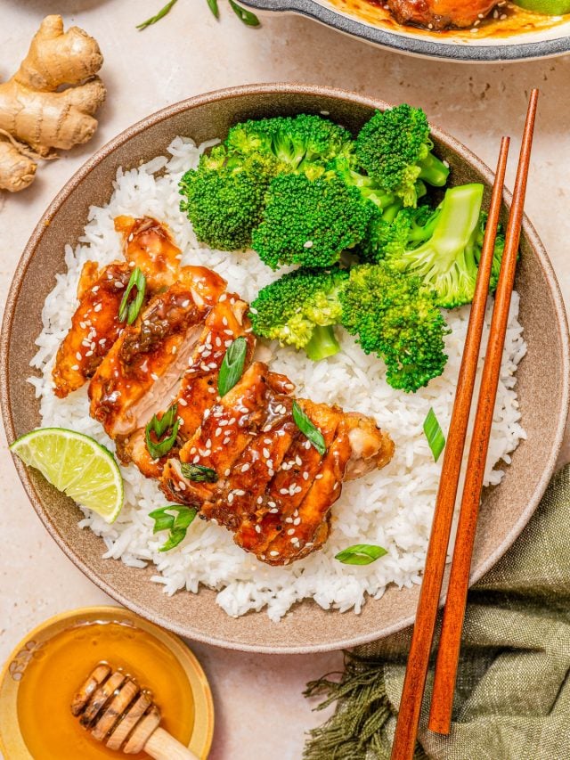 A bowl of white rice topped with glazed chicken, broccoli florets, sliced green onions, sesame seeds, and a lime wedge, with chopsticks resting on the side.