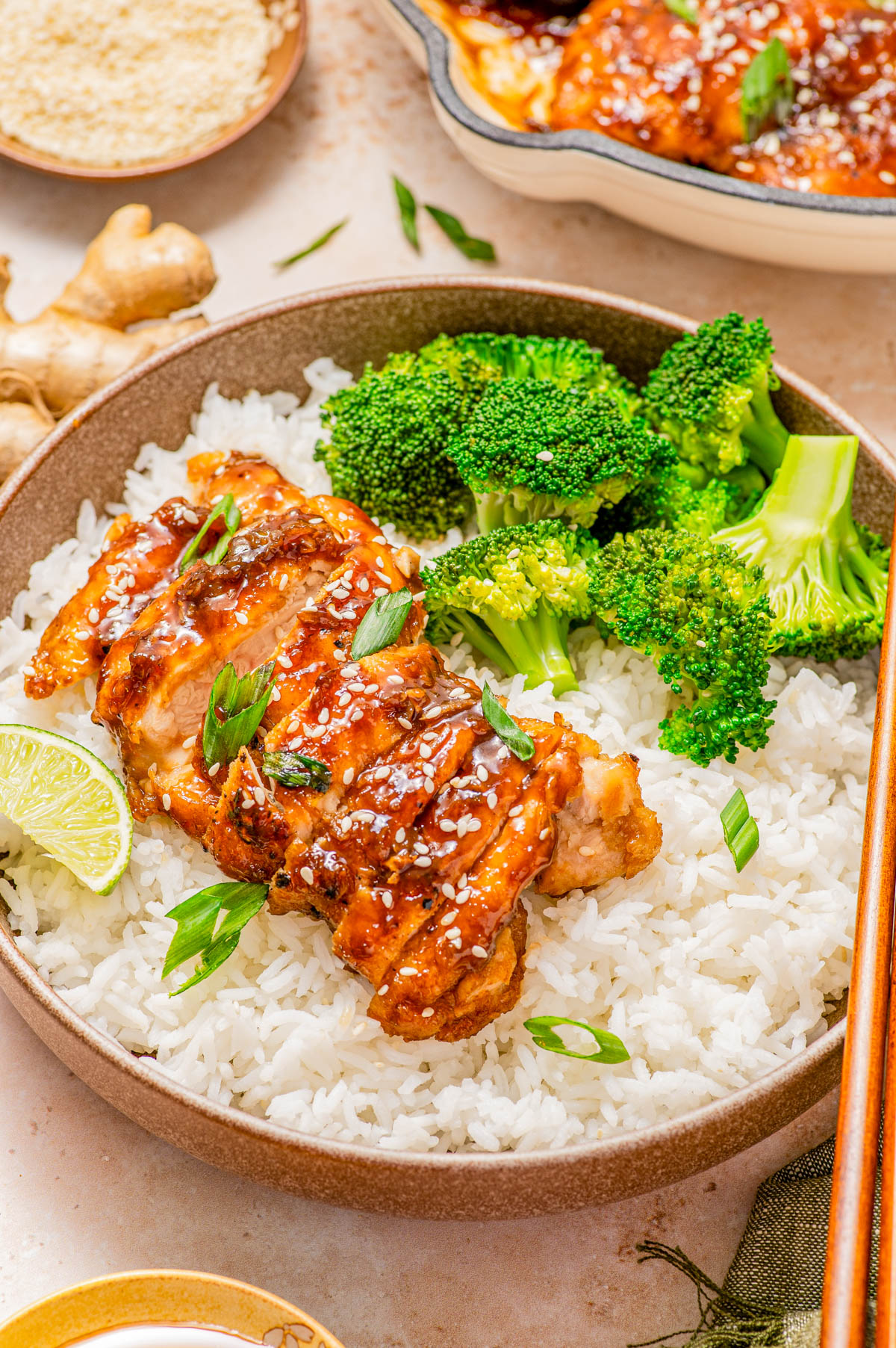 A bowl of white rice topped with glazed chicken, sesame seeds, chopped green onions, steamed broccoli, and a wedge of lime.