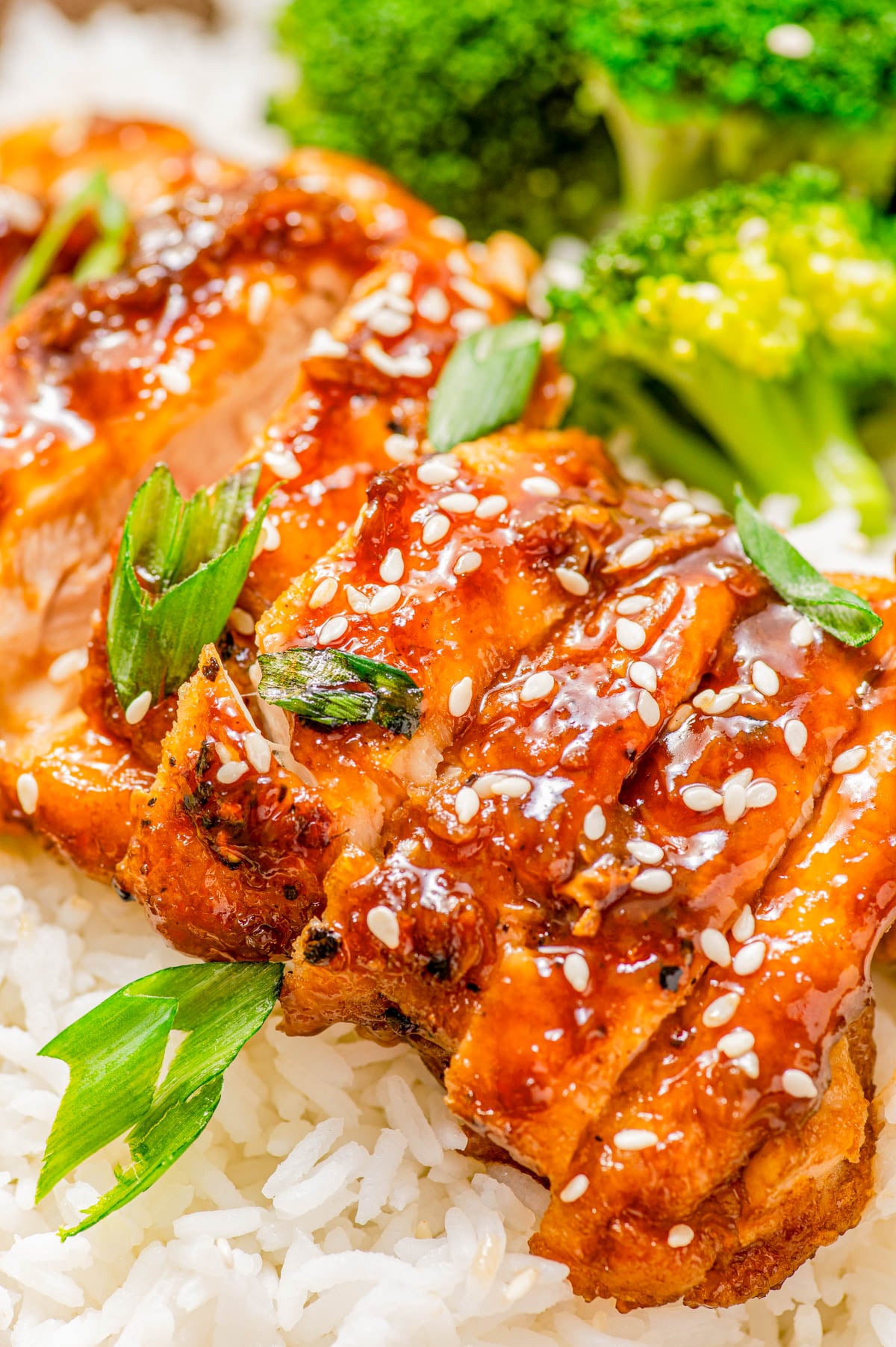 Grilled glazed chicken topped with sesame seeds and green onions, served on white rice with broccoli in the background.