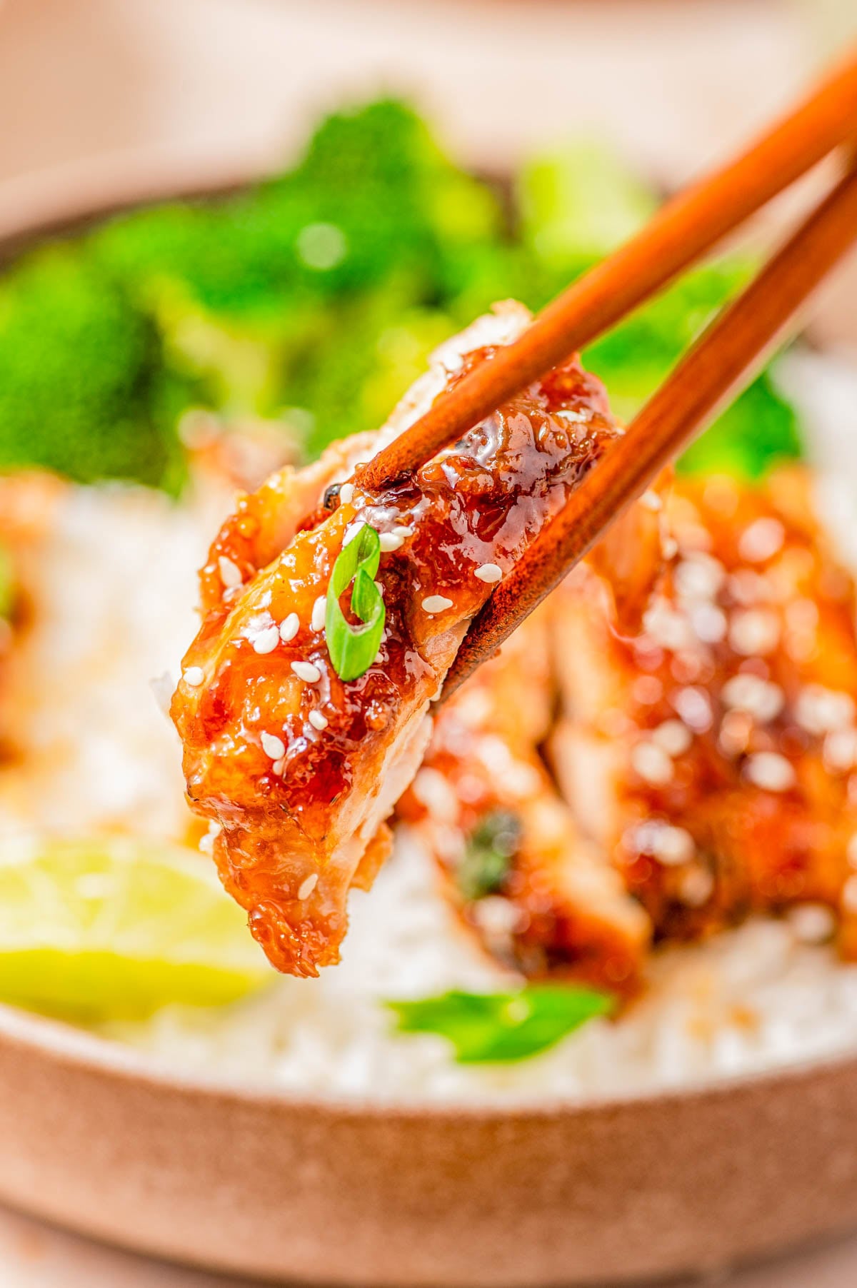 Chopsticks holding a piece of glazed chicken with sesame seeds and green onion, above a bowl of white rice and broccoli.