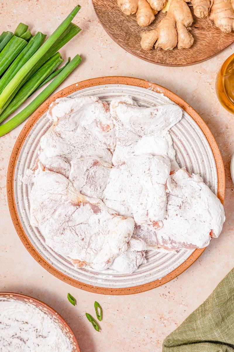 A plate of raw chicken pieces coated in flour sits on a countertop, surrounded by green onions, fresh ginger, a small bowl of oil, and a green napkin.