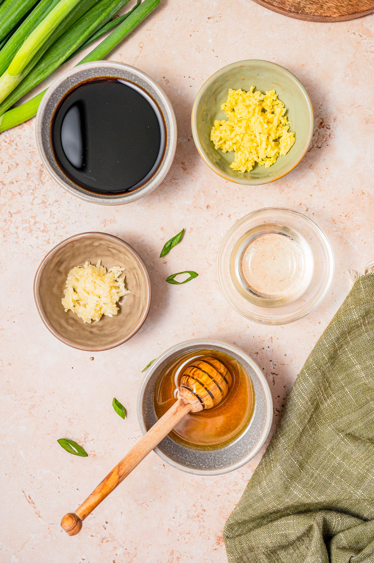 Bowls with soy sauce, minced ginger, minced garlic, rice vinegar, and honey with a dipper are arranged on a light surface next to green onions and a green cloth.