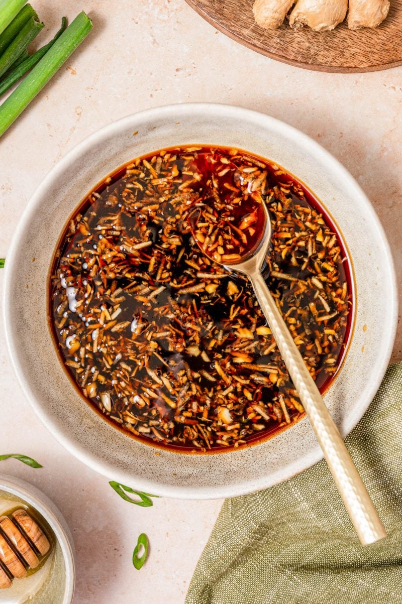 A bowl of shredded ginger and chili oil marinade with a spoon, placed on a light surface next to green onions and a piece of ginger.