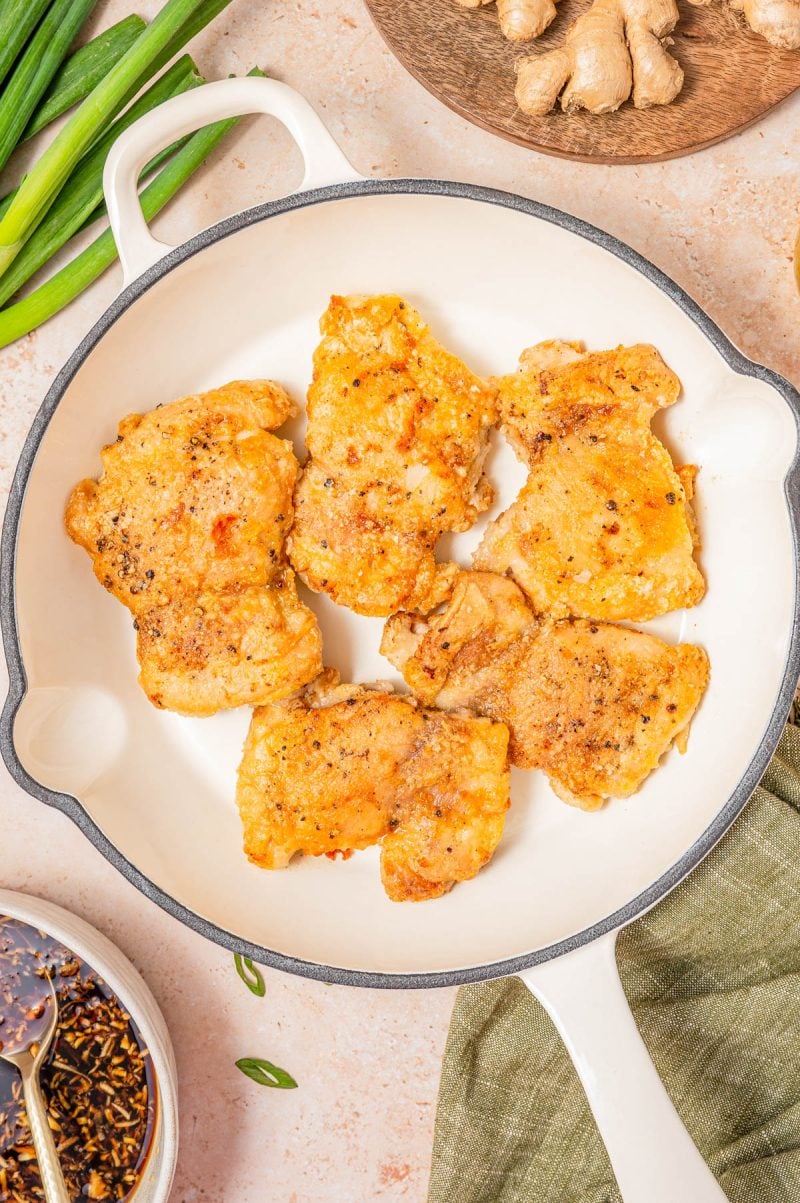 Six pieces of seasoned, cooked chicken thighs in a white skillet, with green onions, ginger, and a bowl of sauce nearby.