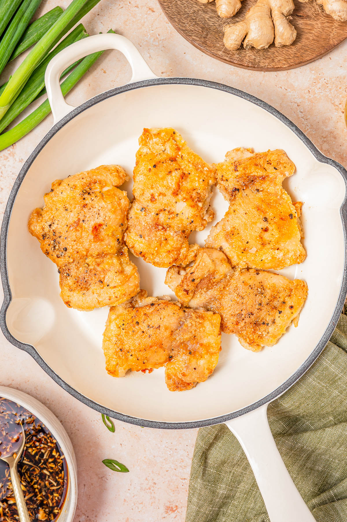 Six pieces of seasoned, cooked chicken thighs in a white skillet, with green onions, ginger, and a bowl of sauce nearby.