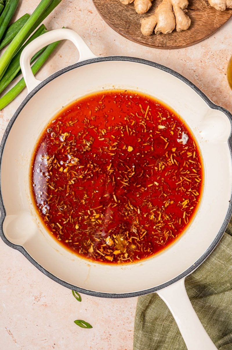 A white pot filled with a red, oily sauce containing visible herbs and spices. Green onions and ginger root are placed nearby on the countertop.