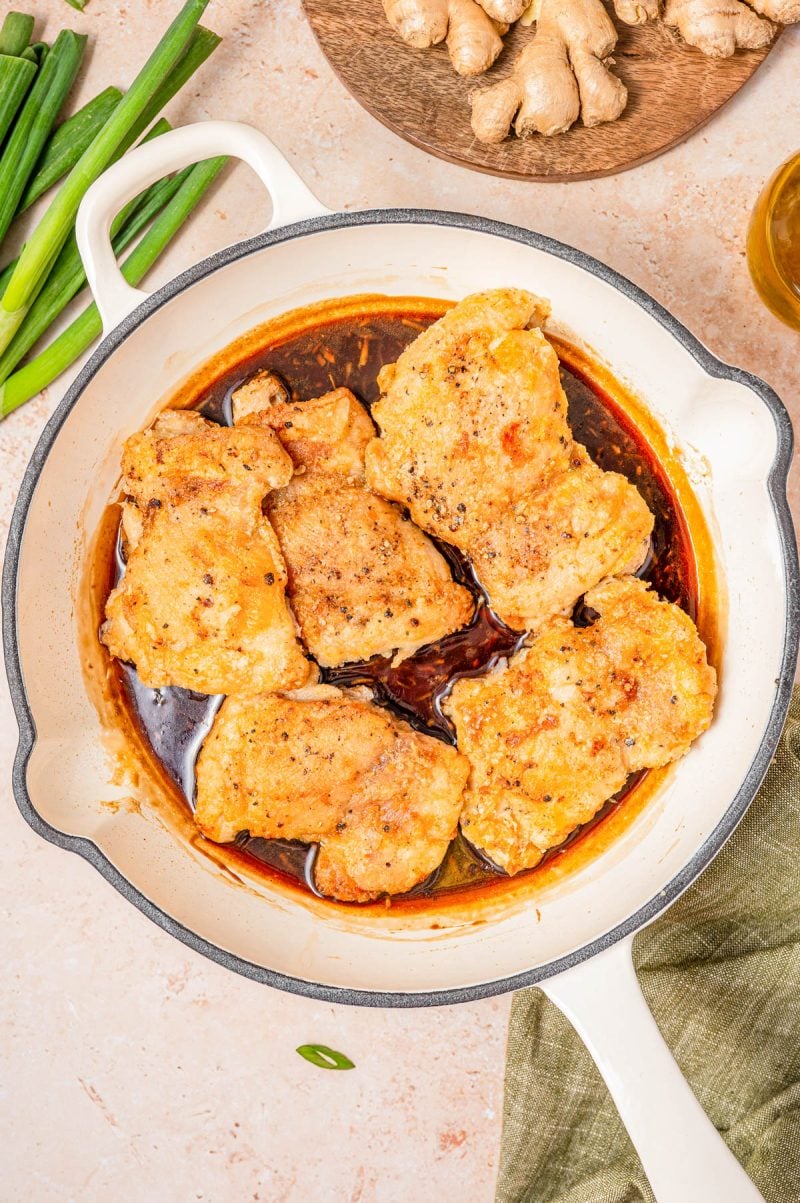 Six pieces of cooked chicken in a white skillet with a brown sauce. Green onions and fresh ginger are nearby on the counter.