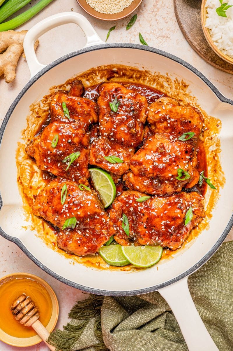 Cooked chicken thighs glazed in a brown sauce, garnished with sesame seeds, sliced green onions, and lime wedges in a round white skillet. Rice and honey are nearby.