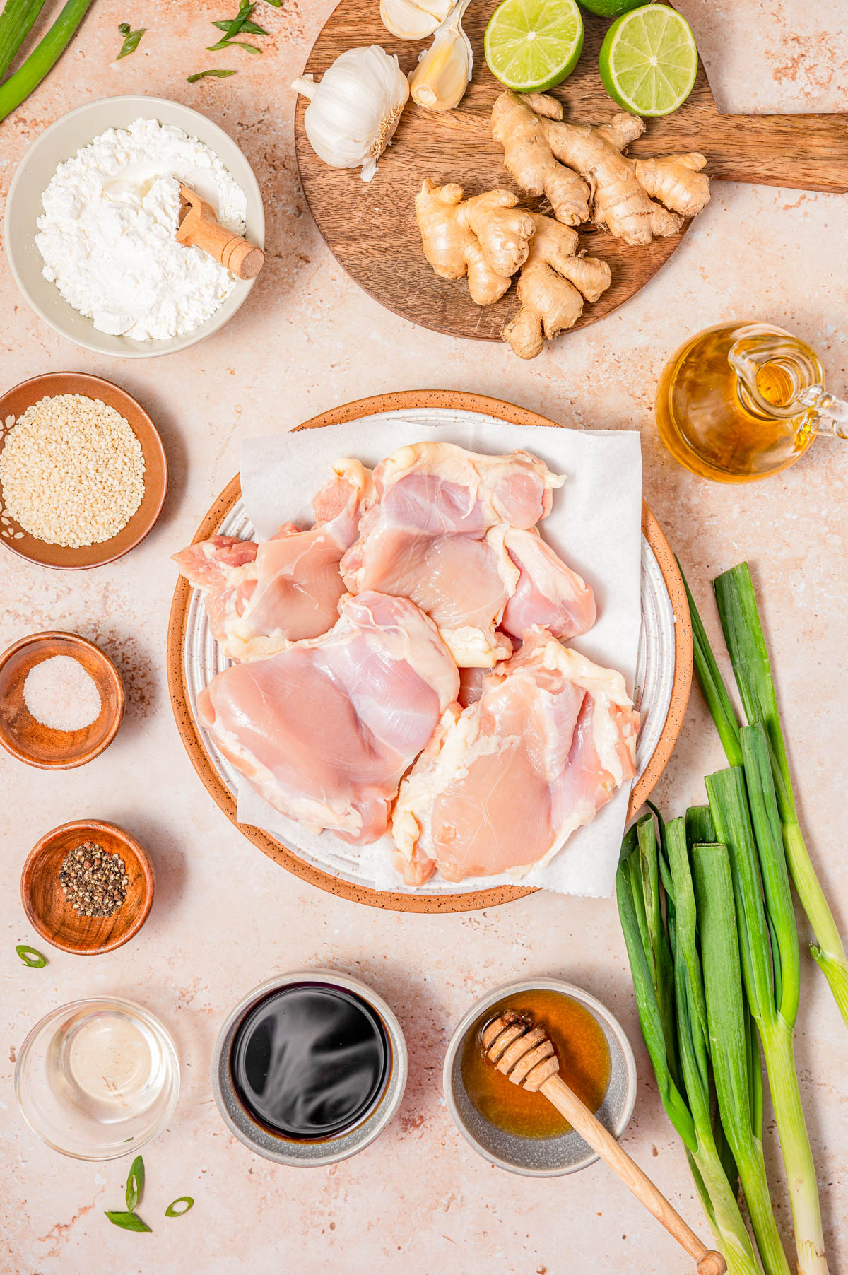 Raw chicken thighs on a plate surrounded by green onions, soy sauce, honey, ginger, garlic, lime, oil, flour, sesame seeds, and seasoning on a light countertop.