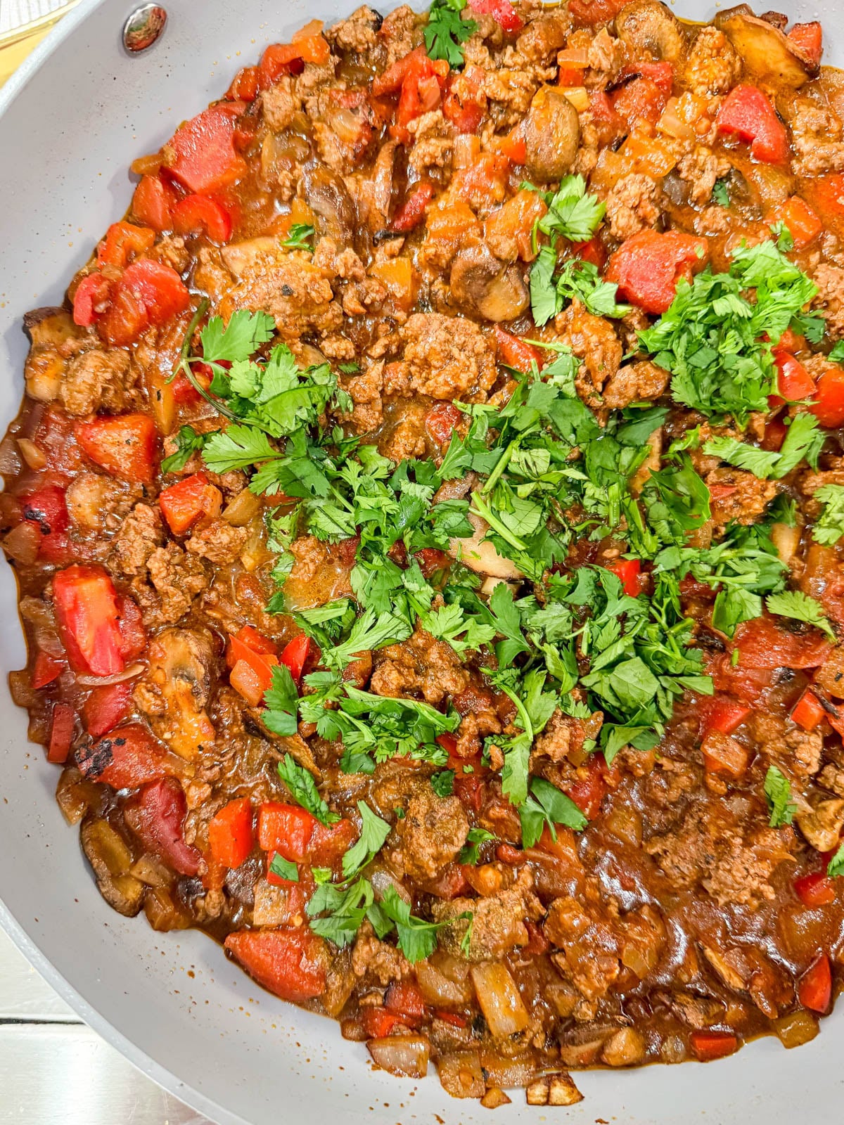 A skillet filled with cooked ground meat, diced tomatoes, onions, red bell peppers, and topped with chopped fresh parsley.
