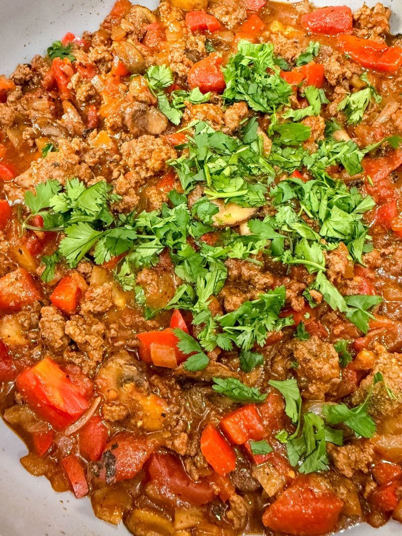 A close-up of a cooked ground meat dish mixed with chopped red bell peppers, onions, and tomatoes, topped with fresh cilantro.