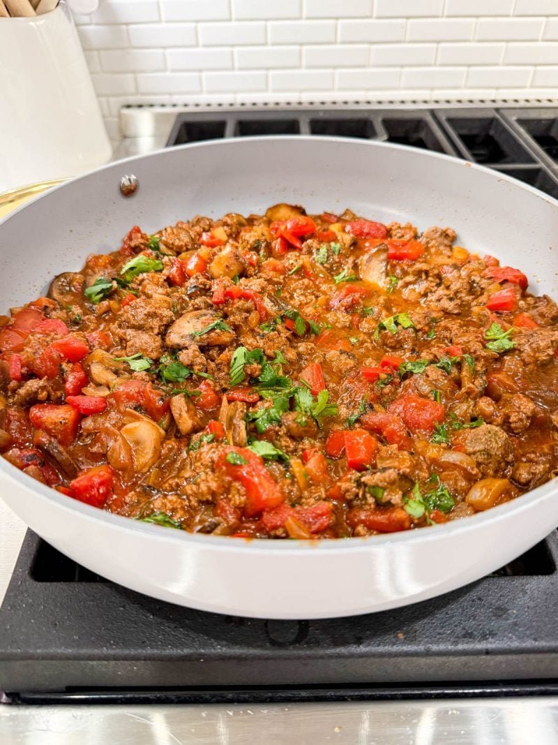 A skillet filled with chunky tomato and ground meat sauce, garnished with fresh herbs, sits on a stovetop in a kitchen with a white tile backsplash.