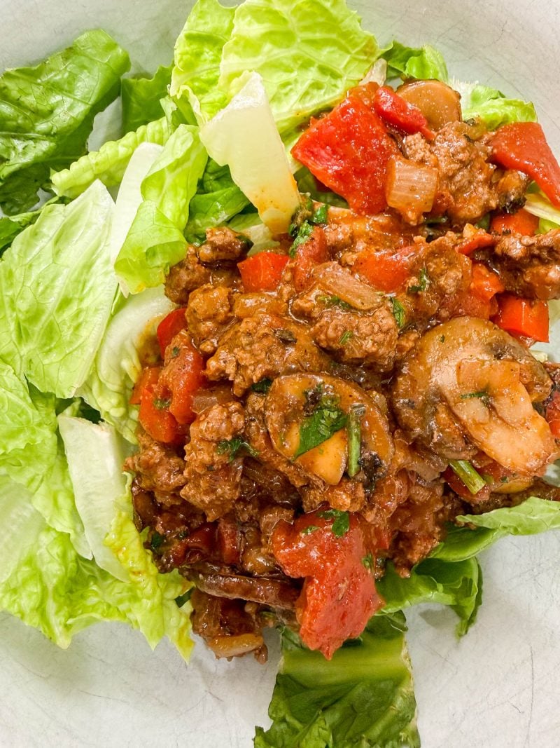A plate of lettuce leaves topped with a mixture of ground meat, tomatoes, red bell peppers, mushrooms, and herbs.
