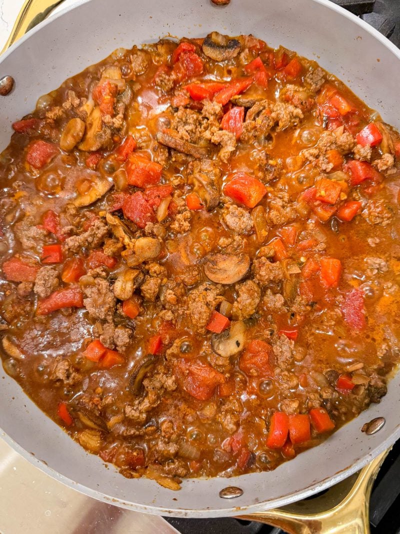 A skillet filled with a cooked mixture of ground beef, tomatoes, mushrooms, and red bell peppers in a red sauce.