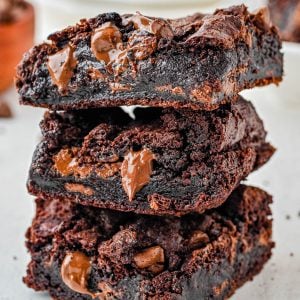Three thick, gooey chocolate brownies with melted chocolate chips are stacked on a light-colored surface.