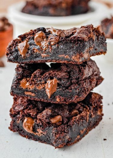 Three thick, gooey chocolate brownies with melted chocolate chips are stacked on a light-colored surface.