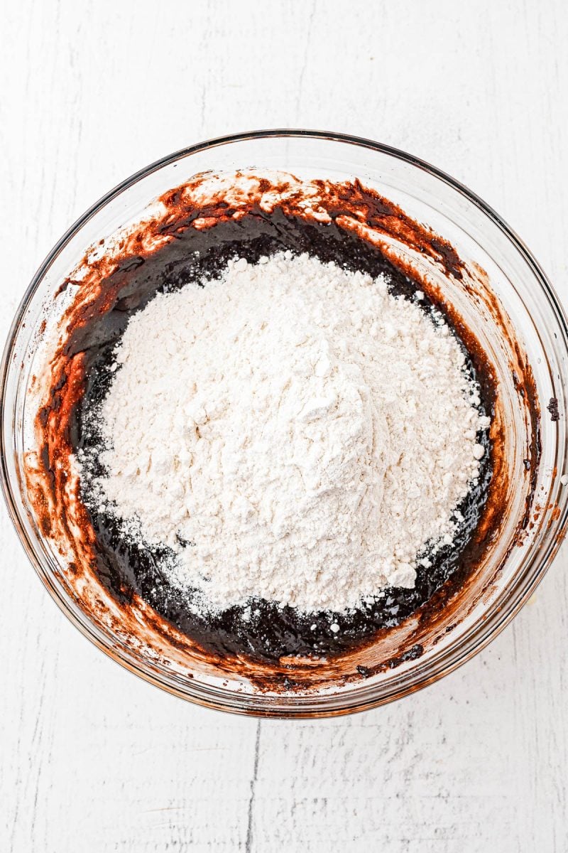 A glass bowl filled with chocolate batter and a mound of flour on top, ready to be mixed, sits on a white surface.