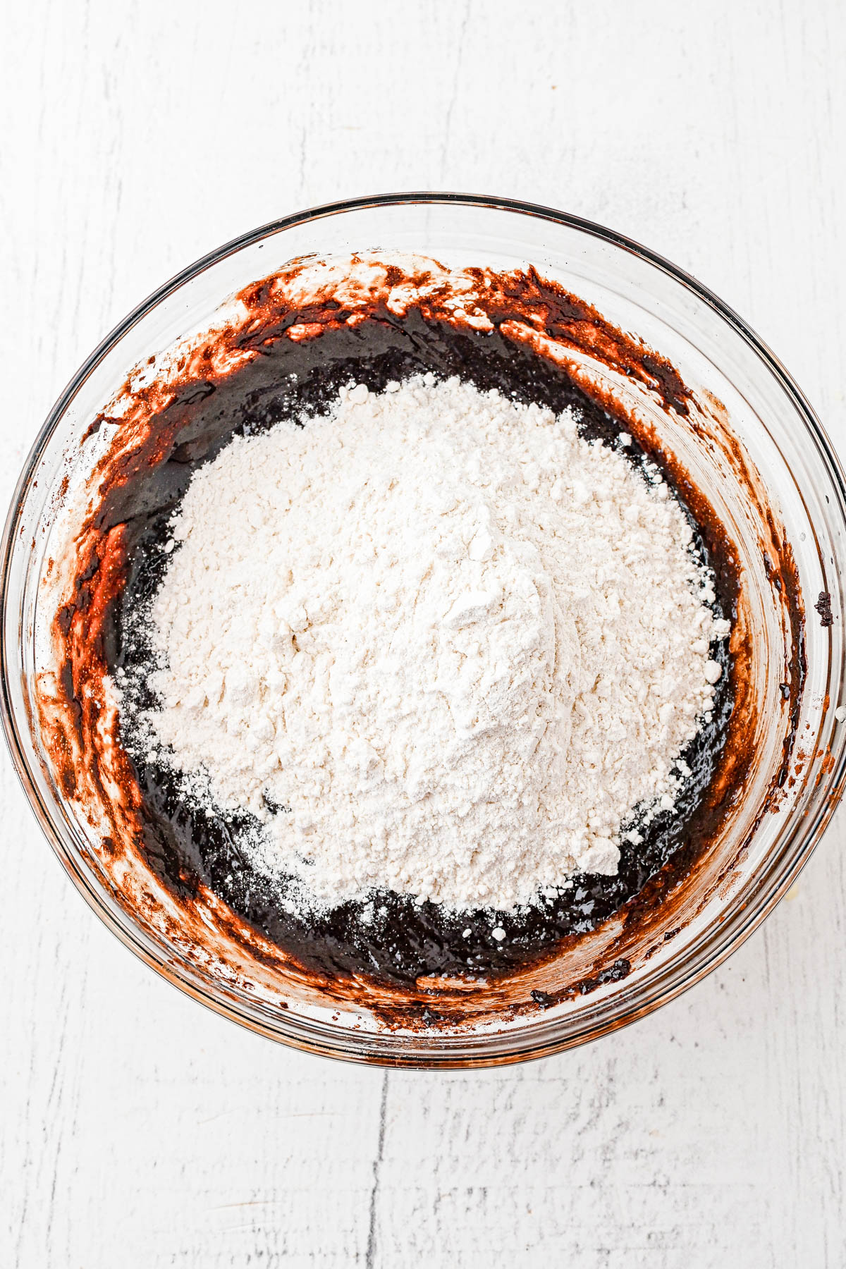 A glass bowl filled with chocolate batter and a mound of flour on top, ready to be mixed, sits on a white surface.