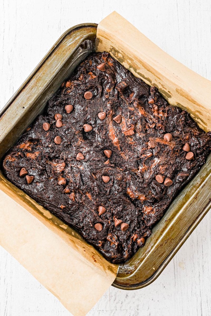 A pan of uncut, baked brownies with chocolate chips on top sits on parchment paper in a square baking dish.