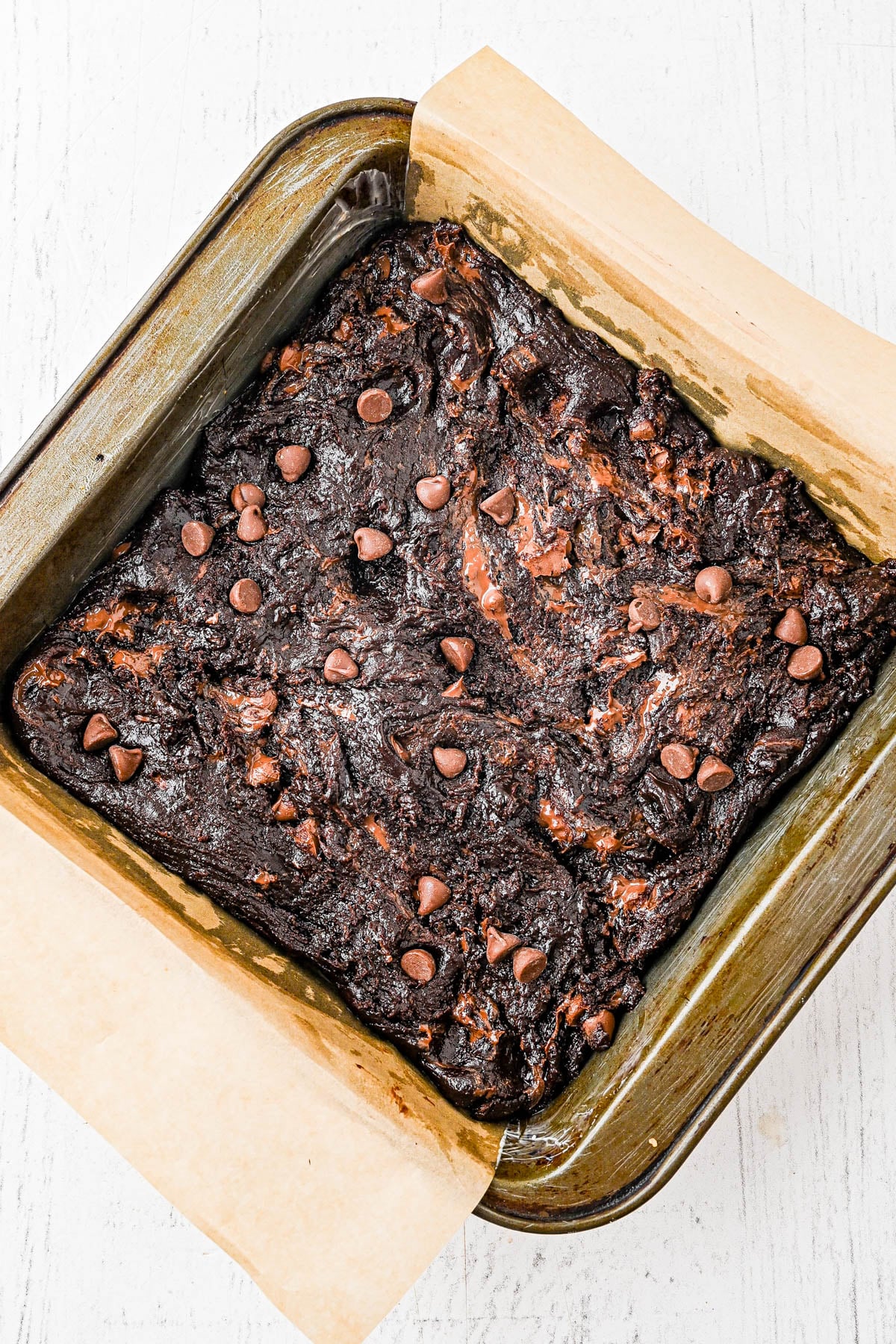 A pan of uncut, baked brownies with chocolate chips on top sits on parchment paper in a square baking dish.
