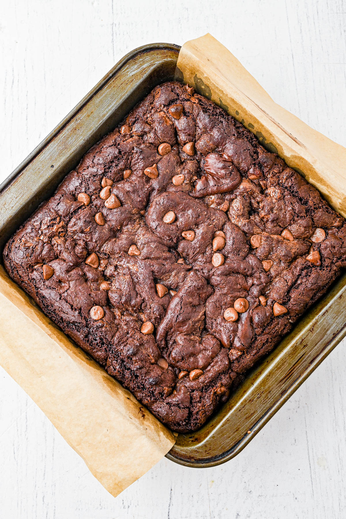 A square baking pan with parchment paper holds freshly baked chocolate brownies topped with chocolate chips on a white surface.