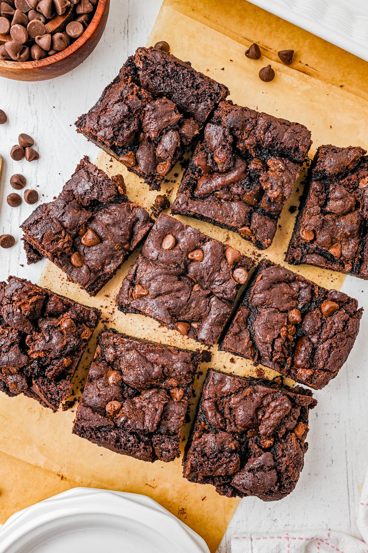 Nine chocolate brownies with chocolate chips are arranged on parchment paper, with some chocolate chips scattered nearby and a white plate partially visible in the corner.