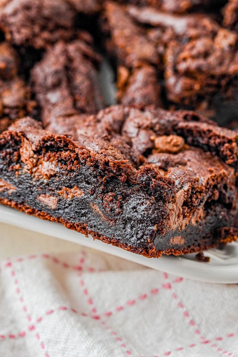 A close-up of a thick, fudgy chocolate brownie with a rich, moist texture, resting on a white plate near a cloth with a pink grid pattern.