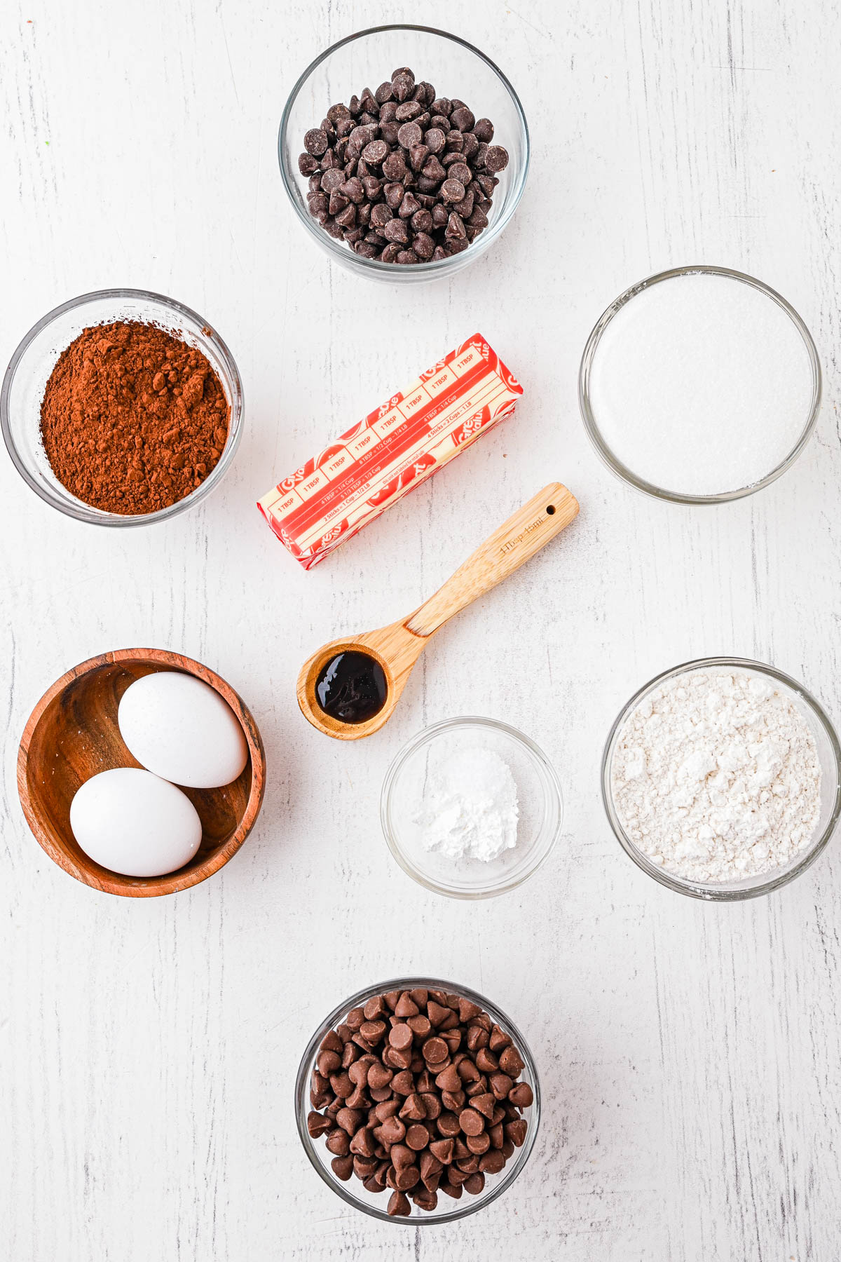 A flat lay of baking ingredients: two eggs, flour, sugar, cocoa powder, two bowls of chocolate chips, baking powder, vanilla extract, and a stick of butter on a white surface.