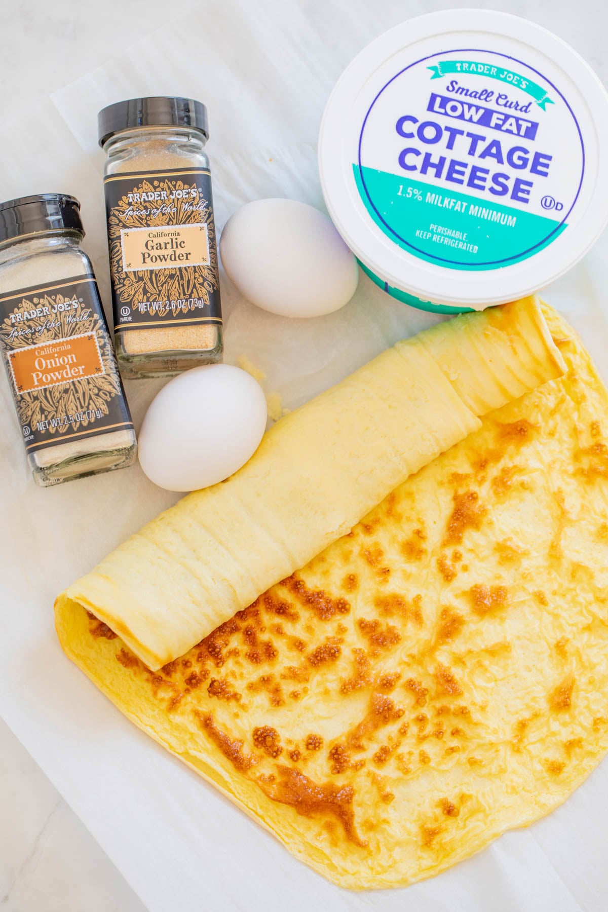 A rolled cooked egg sheet, two raw eggs, onion powder, garlic powder, and a container of low fat cottage cheese are arranged on a white surface.