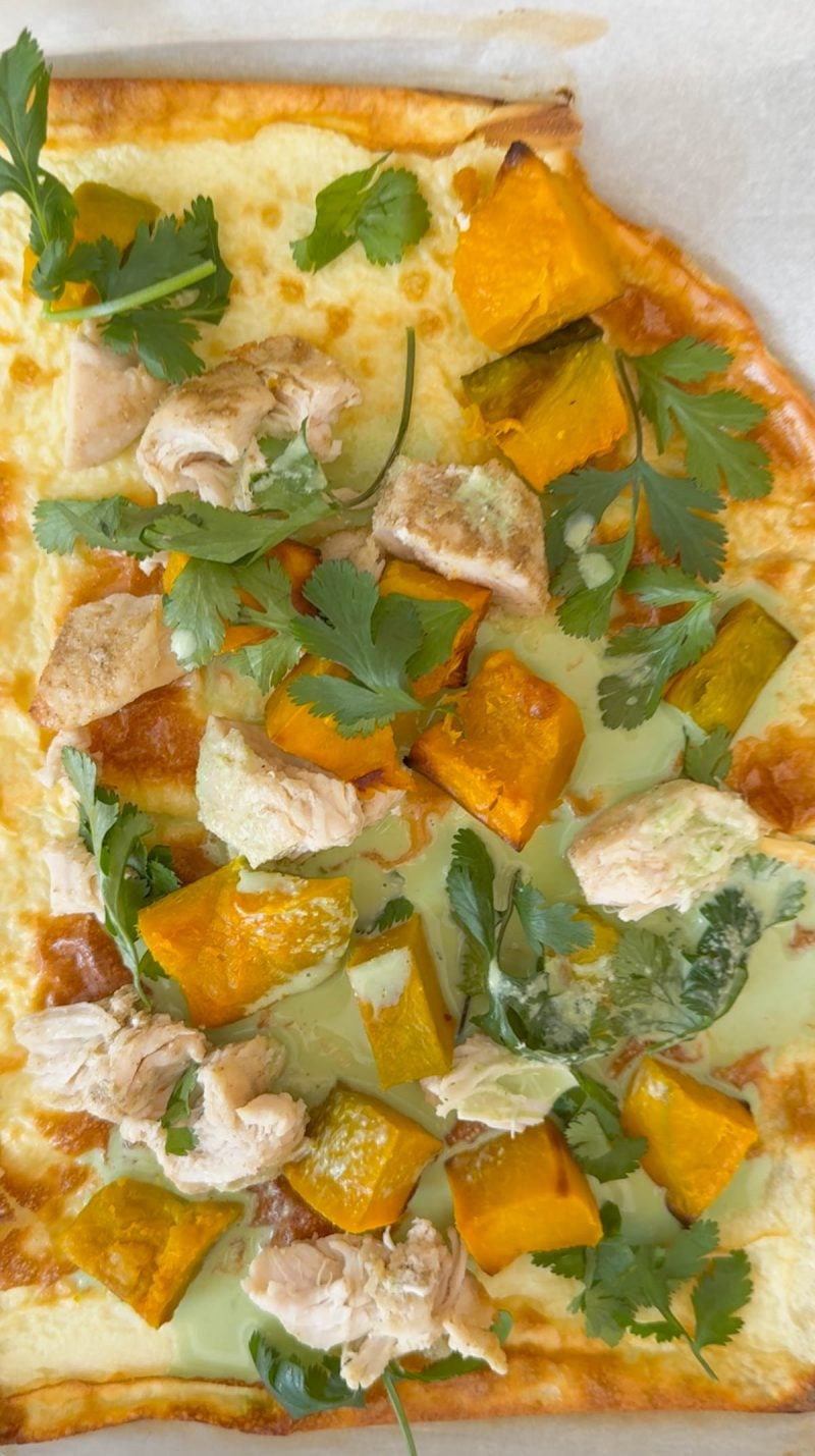 A close-up of a flatbread topped with roasted pumpkin cubes, pieces of chicken, fresh cilantro leaves, and a drizzle of green sauce on parchment paper.