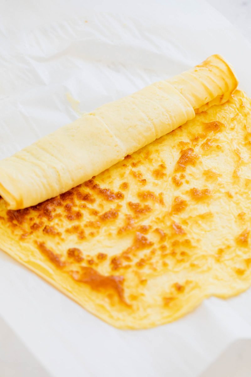 A thin, yellow baked egg sheet partially rolled up on a sheet of white parchment paper.