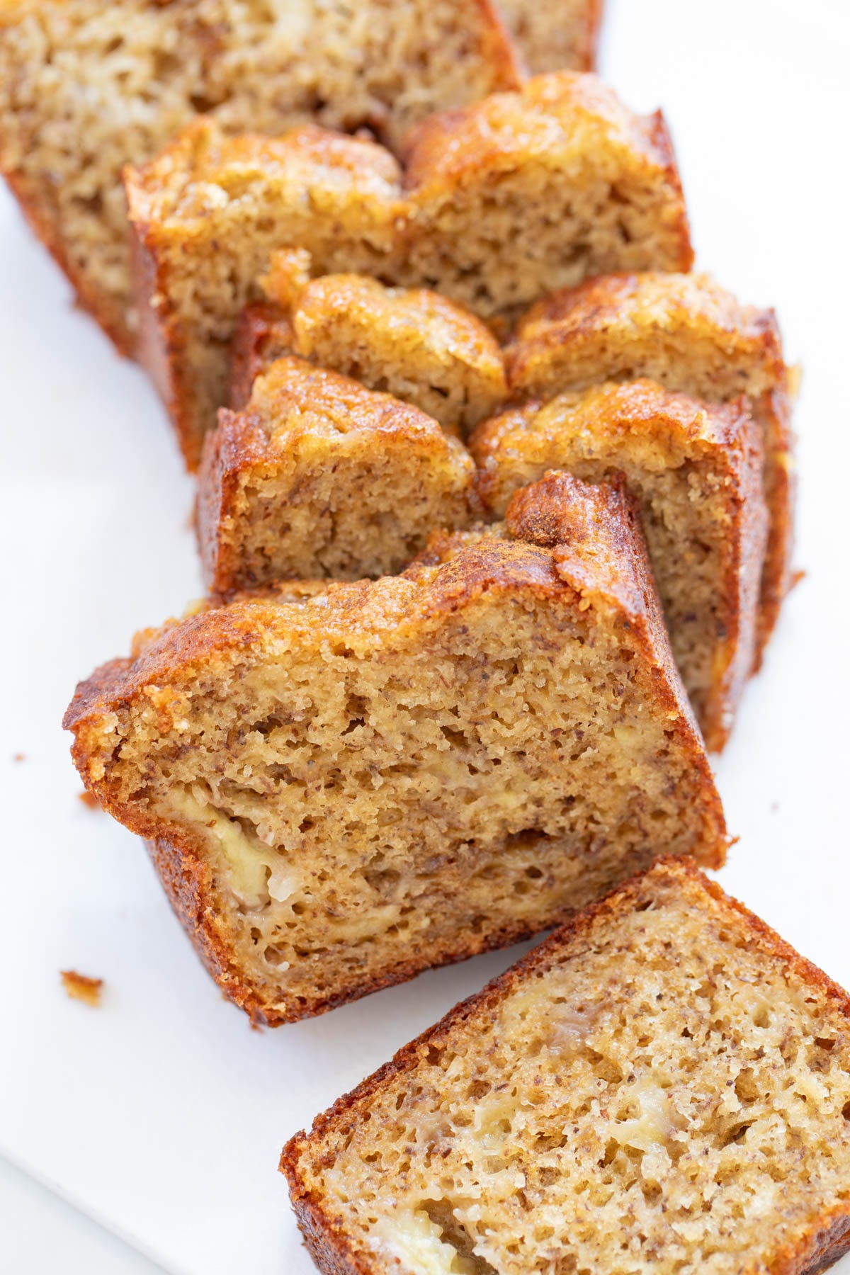 Sliced banana bread on a white surface, showing a moist and textured interior with a golden-brown crust.