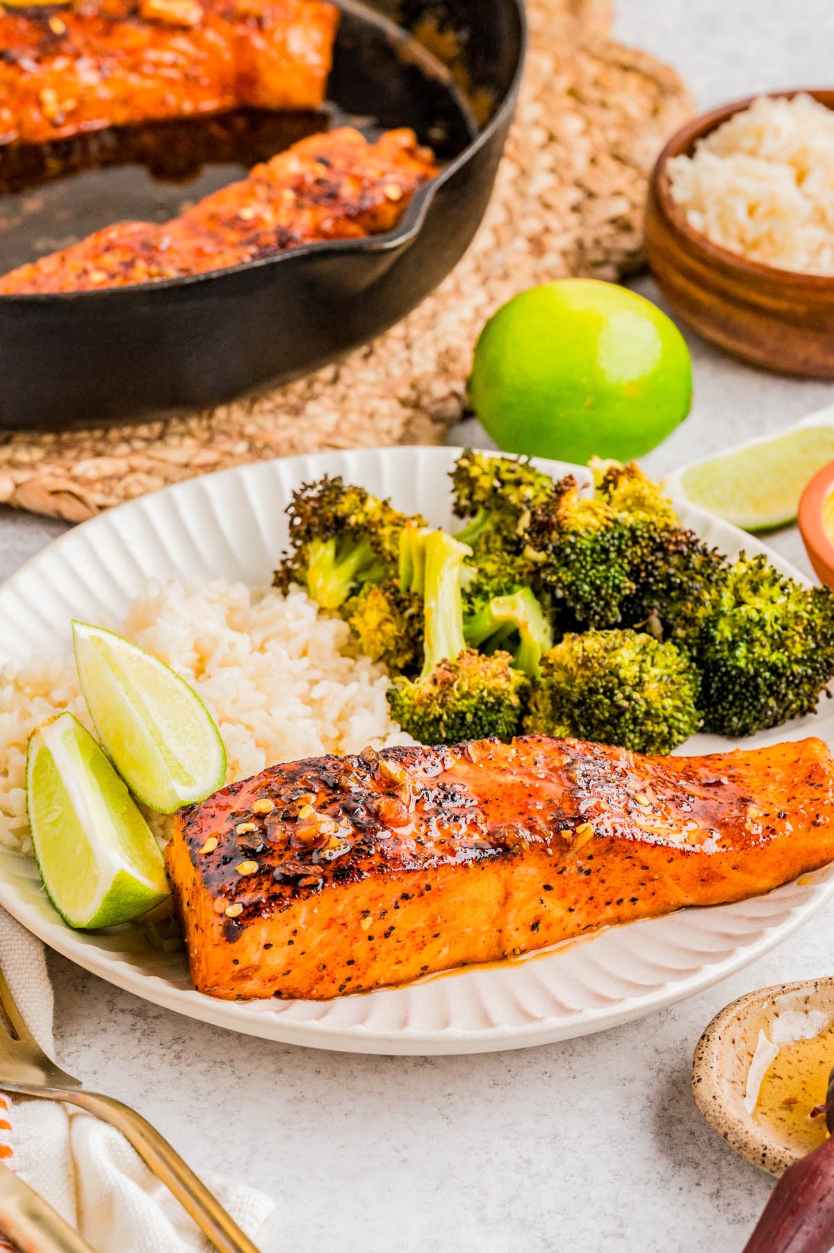 A plate with a cooked salmon fillet, white rice, roasted broccoli, and lime wedges, with a skillet, bowl of rice, and lime in the background.