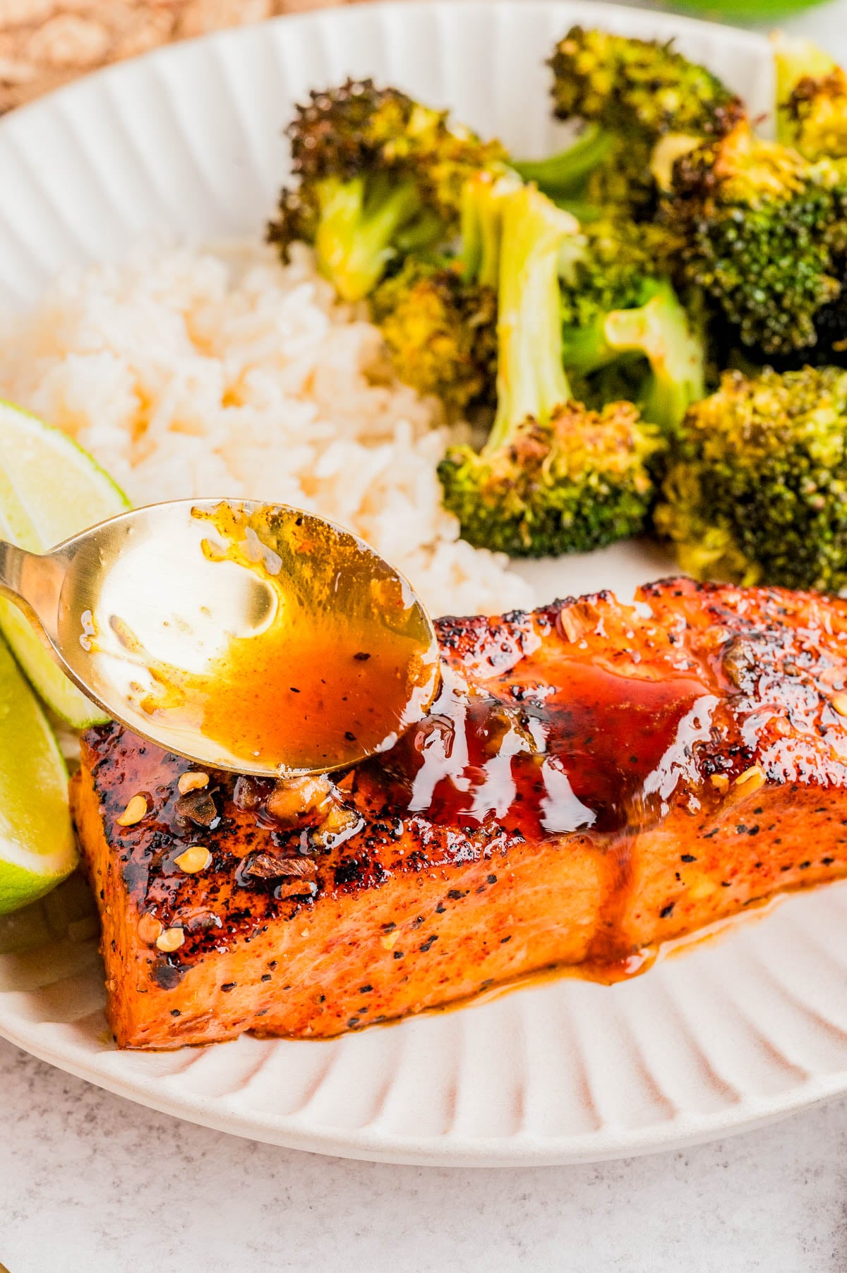 A spoon drizzles sauce over a seasoned salmon fillet on a plate with rice, roasted broccoli, and lime wedges.