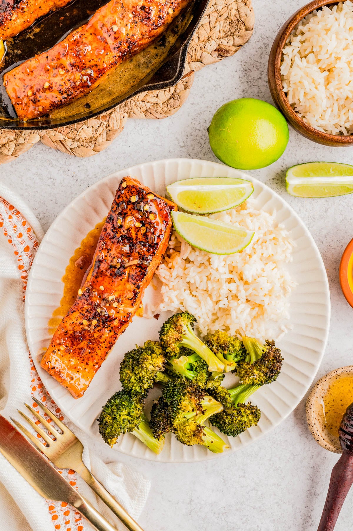 A plate with grilled salmon topped with spices, steamed white rice, roasted broccoli, and lime wedges, with extra rice and a cast iron pan of salmon nearby.