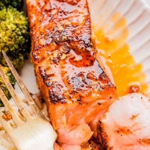 Close-up of a cooked salmon fillet with seasoning and sauce, served with white rice and broccoli on a plate, alongside a fork.
