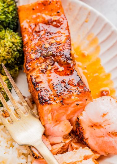 Close-up of a cooked salmon fillet with seasoning and sauce, served with white rice and broccoli on a plate, alongside a fork.