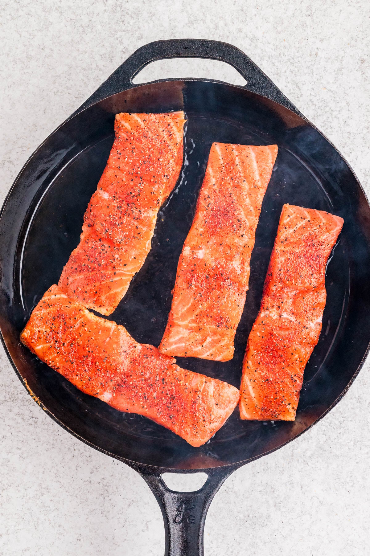 Four seasoned salmon fillets cooking in a black cast iron skillet on a light-colored countertop.